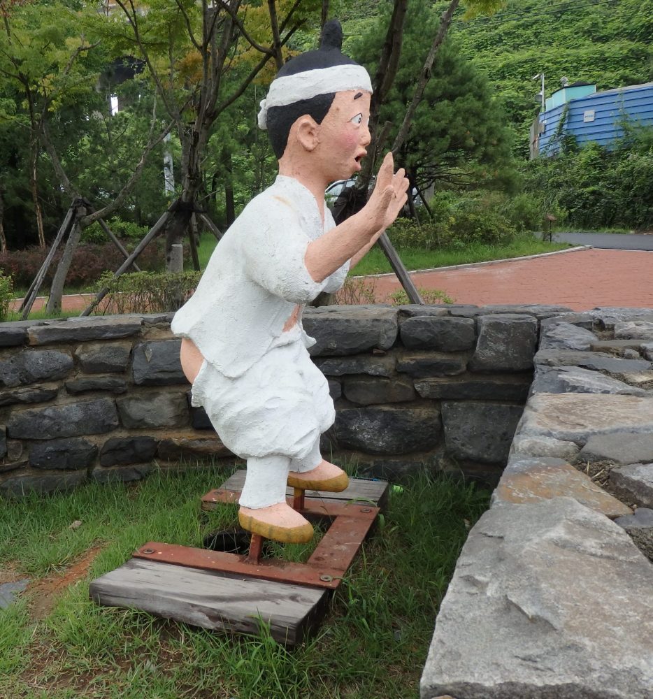 Plaster statue of a Korean peasant slightly squatting, with a surprised look on his face, above two stones. He is wearing a white outfit and has a white headband tied around his head. His pants are pulled down around his thighs and his hands are up in the air as if someone is pointing a gun at him. 