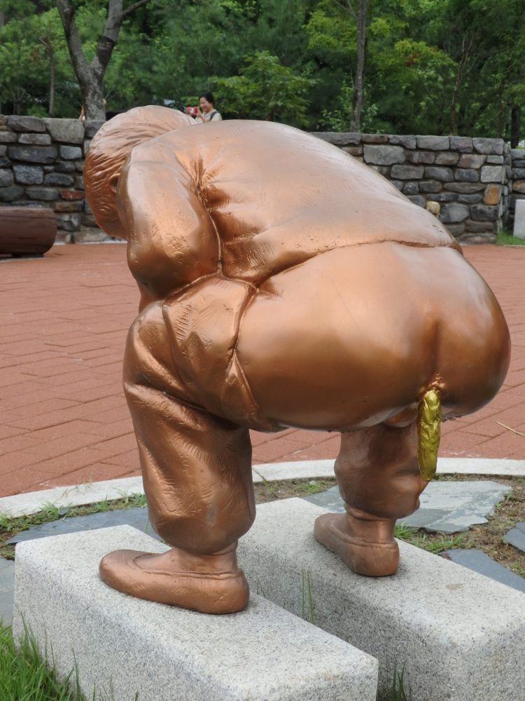 The statue is in a copper color: a figure of a man, standing on a historical toilet consisting of two rectangular slabs of stone. The idea is to stand on the stones and poop/pee between them. In the photo the man is facing away from the camera with his pants pulled down to mid-thigh. He is squatting and a large poop is emerging from his anus, hanging down. The poop is for some reason, gold-colored. 