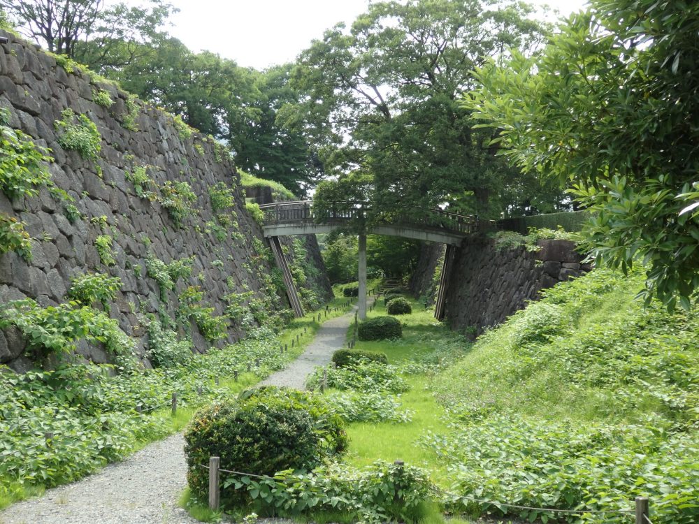 The moat has a grassy floor with a path running through it. On the left a steep, almost vertical wall of large stones. ON the right a grass-covered wall that is lower. A foodbridge connects the tops of both walls, crossing the path underneath.