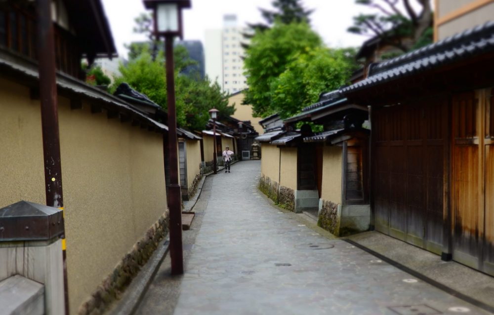 a street in the Nagamachi neighborhood of Kanazawa: low buildings on either side with light brown plastered walls and brown roofs. No windows are visible. The road curves out of sight and a tall modern building is visible in the distance. Is Kanazawa worth a visit?