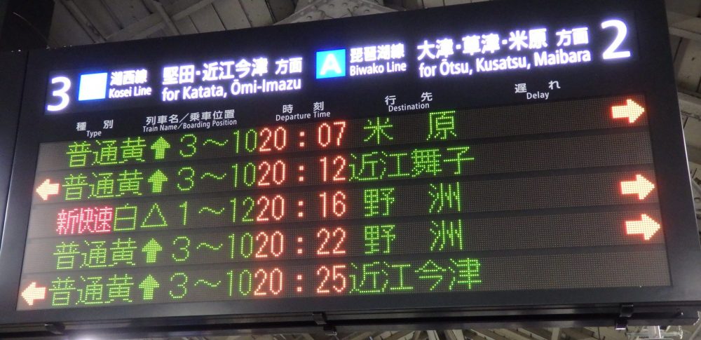 an electronic sign lists 5 trains, some arriving on the left-hand platform (marked as track 3 for Katata and Omi-Imazu) , some on the right (track 2 for Otsu, Kusatsu and Maibara). Next to 4 of them are upward arrows. ONe shows a triangle.