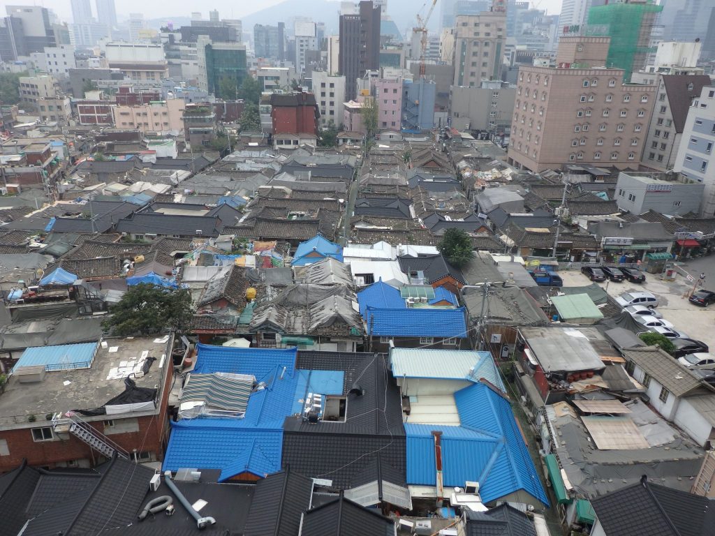 view of Ikseon Hanok Area from my hotel room shows low roofs in various conditions, some newly roofed in blue but most gray with patching. Lots of taller buildings in the background.