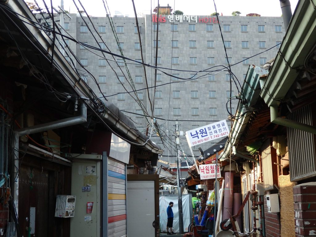 In the background, the Ibis Hotel looms, a flat gray building with many stories, over the Ikseon Hanok Area houses in the foreground: single-story houses along a very narrow street witha mess of electric cables over it.