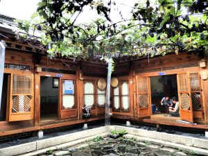 A hanok interior courtyard in Buckchon Hanok Village - wooden filigreed doors open out to the courtyard, which has a small pond with a vine shading it. Two men work in one of the doorways, their shoes lined up outside the door on the ground.