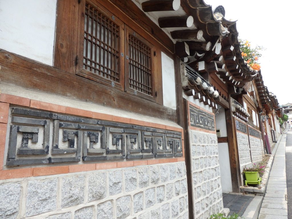 exterior of a hanok in Buckchon - stone below, decorative brick work above, then above that, wood beams, windows, white plaster, and above that round wooden roof beams sticking out.