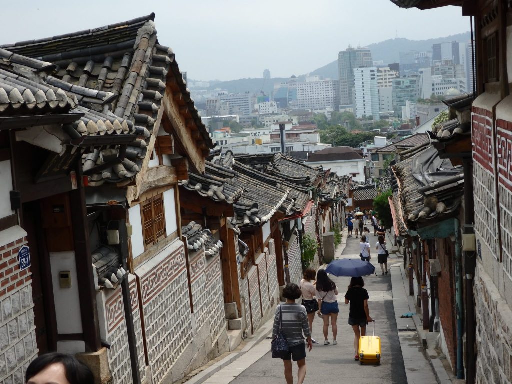 view down a Buckchon Hanok Village street: the street sloped down and the houses on either side are neatly faced with brick and tile and have neat sloping wood and tile roofs.