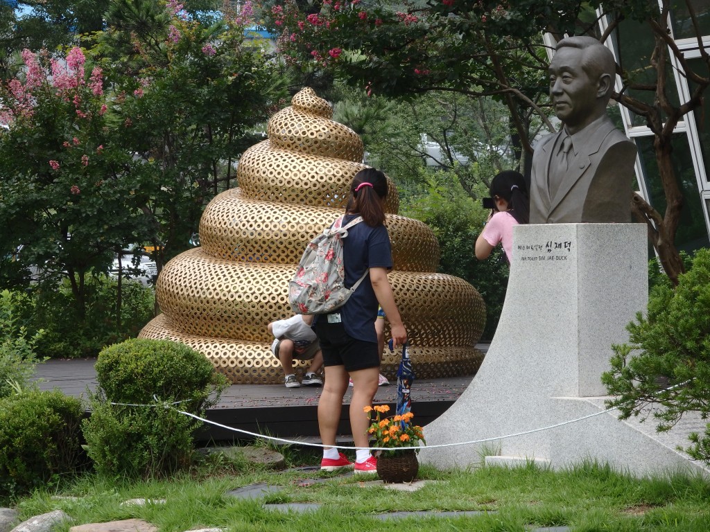 The huge gold turd is about 2 meters high. Two children, partly blocked by a parent, are squatting in front of it, posing for a picture.