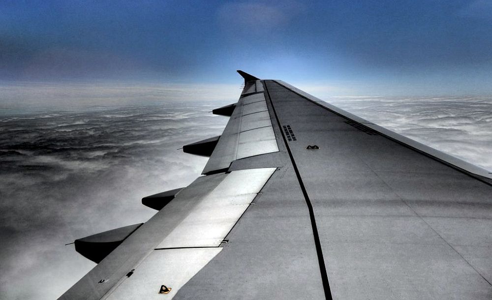 Looking out of a plane along the grey wing. Blue sky in the background, a cloudy sky below.