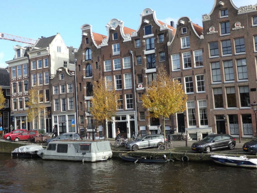 A row of typical Dutch row houses: tall and narrow, most of them are three windows wide and five or six stories high with pretty decorative gables at the top. In the foreground, the water of a canal and a few boats moored along its side.