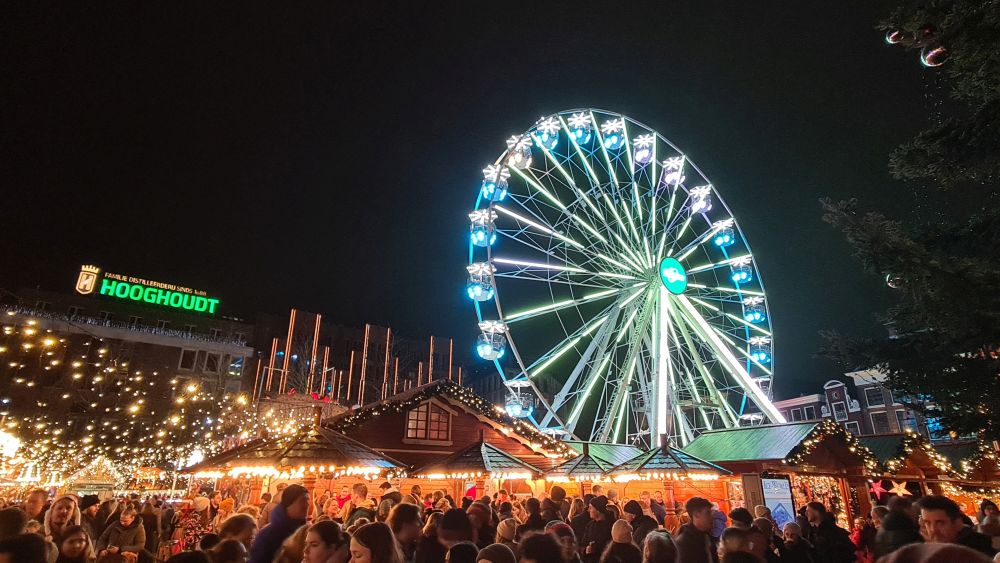 Crowds of people with brightly-lit "chalets" behind them, and a brightly Ferris wheel behind the chalets.