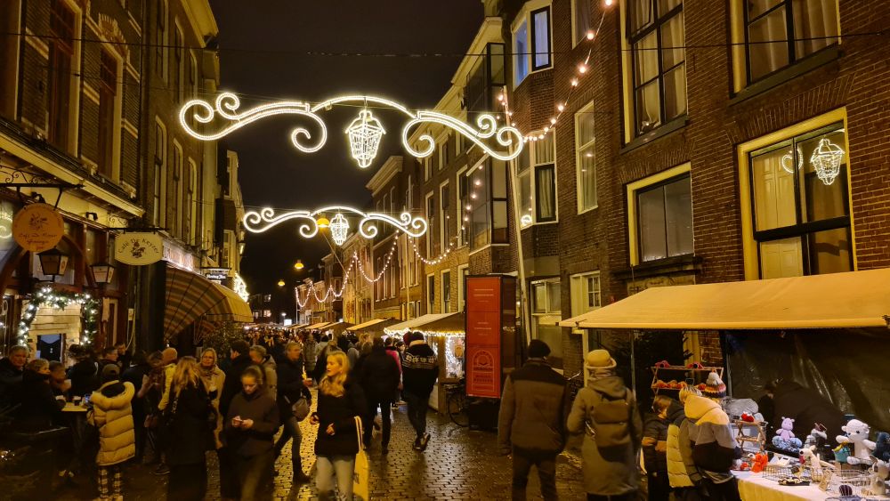 A narrow street with houses on both sides, people walking down the center, and here and there a market stall on either side. Christmas lights suspended across the street.