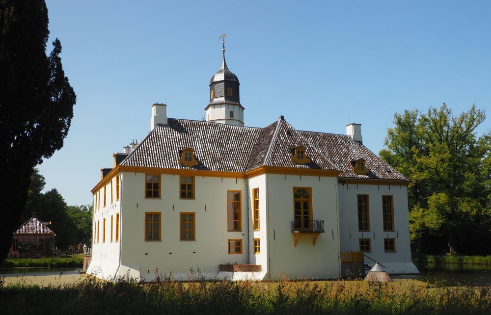 View of the estate house from behind: white-painted with dark yellow around the windows, it's more or less symmetrical, with a section that sticks out more than the rest in the center, and the tower showing behind it.