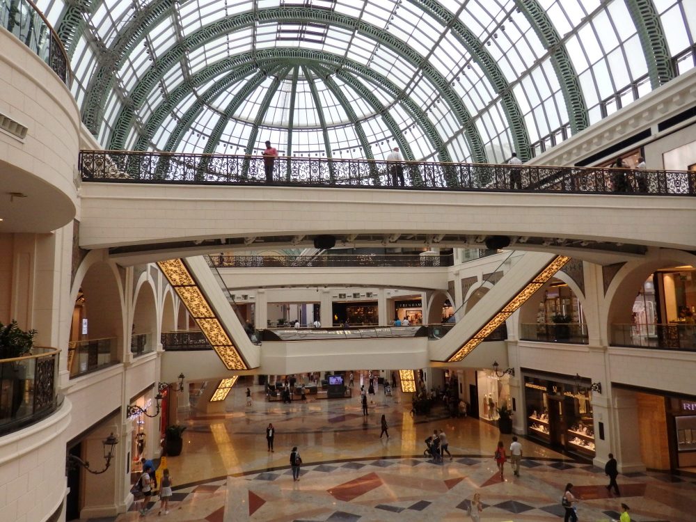The marble floor with scattered people is visible below. Straight ahead and a bit above camera level are a series of bridges and escalators connecting them. The roof above is a half-cylinder and entirely glass panes with a metal framework. 