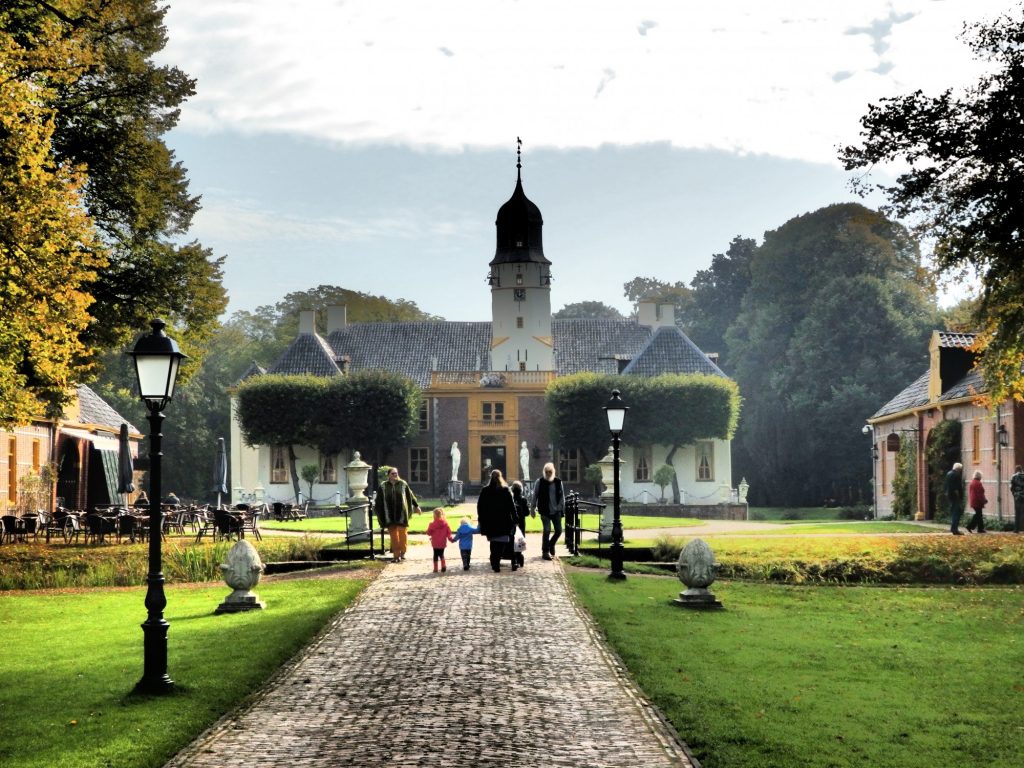 Fraeylemaborg, with the restaurant on the left and coach house on the right. The photo sights straight down the approaching brick walk toward the entrance to the main house. The main house is low and white, with a row of carefuly pruned trees hiding much of the facade. A tower rises behind them, off center from the central entrance.