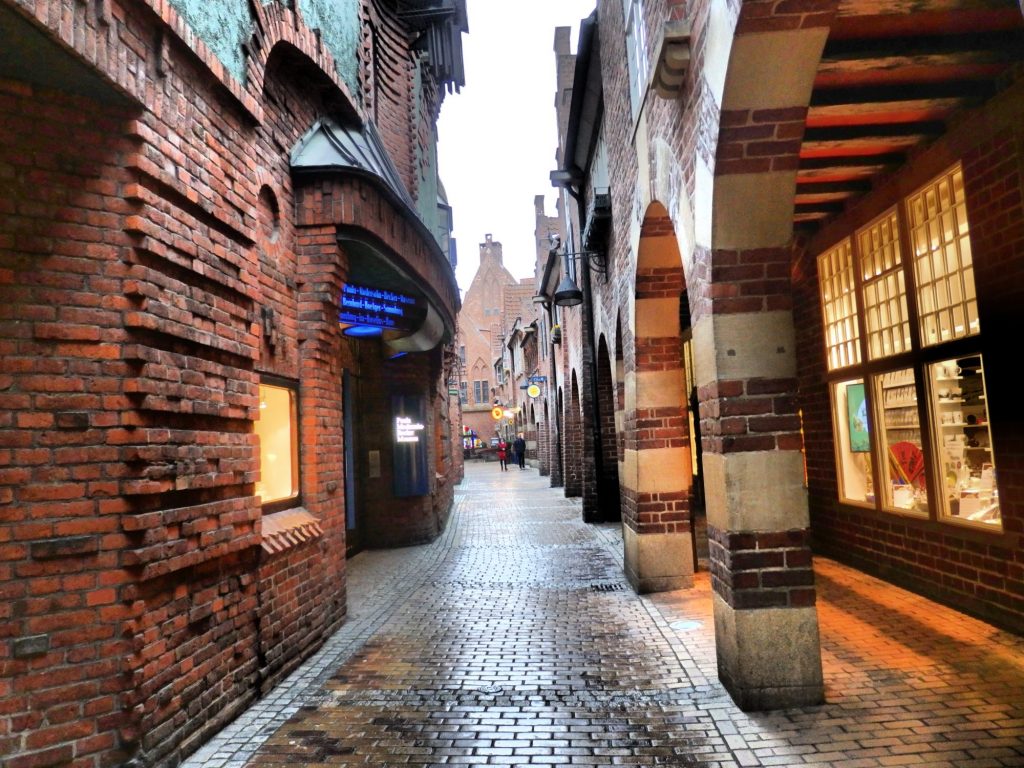 On the right is an arcade with arched columns in alternating red brick and concrete. ON the right is a red-brick building with interesting patters to the brick: sometimes protruding, sometimes not, forming circles and arches in the facade. The street ahead is brick paved.