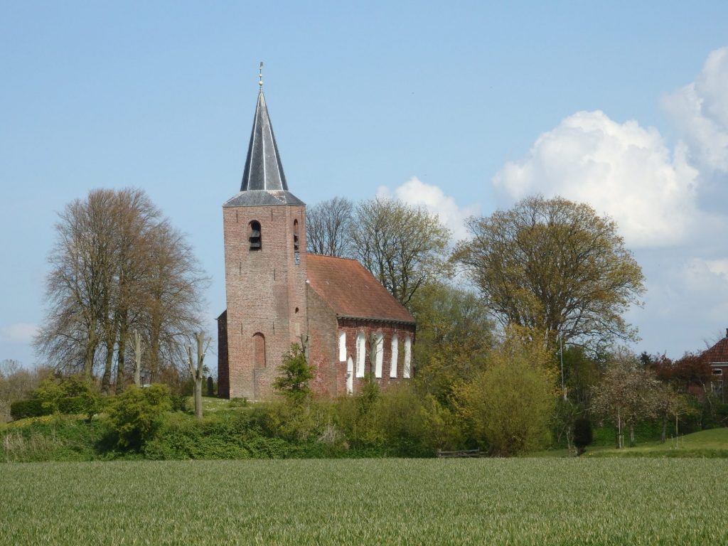 A very small, very simple red-brick church: rectangular, white-edged windows on the side, standing on a slight rise, with a square tower a bit taller than the building in front. The tower has a black pointed roof and leans slightly to the left. In the foreground, a grassy field. A few trees dot the rise (terp) around the church.