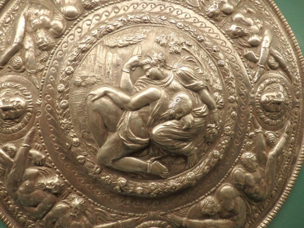 A gold or perhaps bronze shield with a man and woman in the center, his face in her lap. More men around the edges.