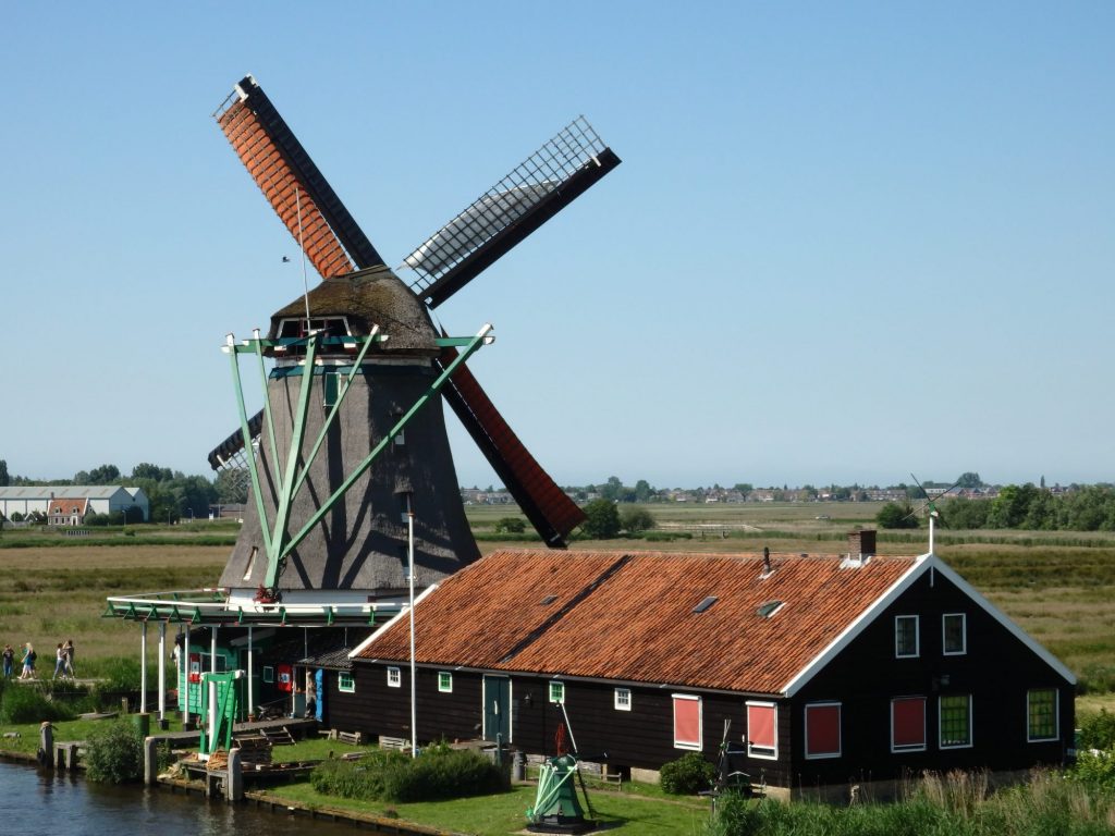 The windmill has an octagonal shape, wider at the bottom than the top. It is covered with thatch. On the back (from this view) are the four windvanes and their sails are unfurled on the windvanes. In front of it is a long low brown building with a low peaked red roof.