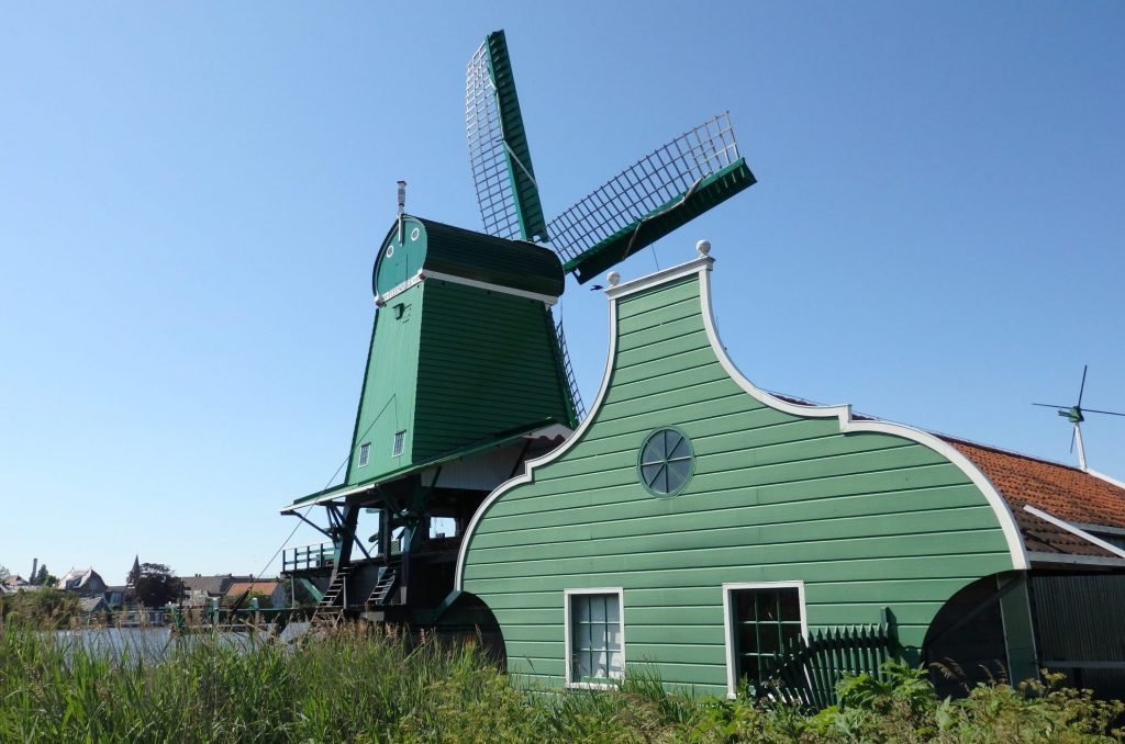 The windmill stands on stilts and is painted green, with a 4-sided body, slightly wider at the base than the roof. A low building next to it has a gable that rises on either side and is flat on the top. It is also made of wood and painted green.