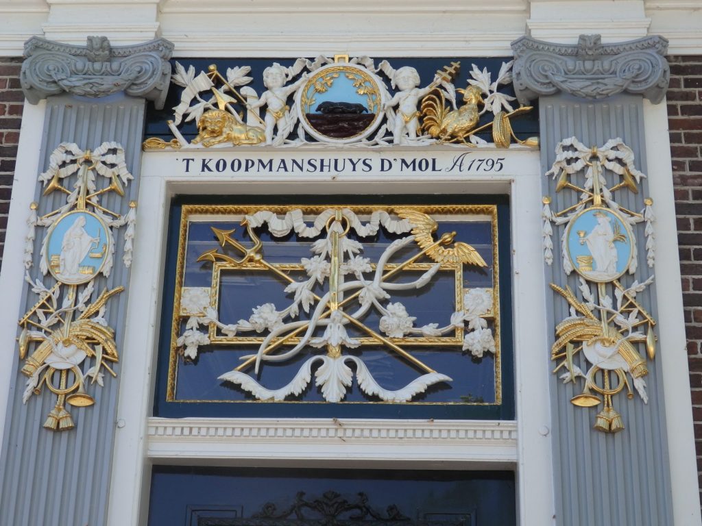 In the square above the door as well as above and beside it are some lavish carvings: feathers, arrows, banners of all sorts, mostly in white or gold. Above the door frame it reads "'t Koopmanshuys d'Mol A1795."