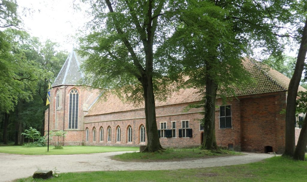 A low, long, red-brick building with what looks like a church attached at one end: it has tall arched windows. In front of the building is a grassy area with some large trees.