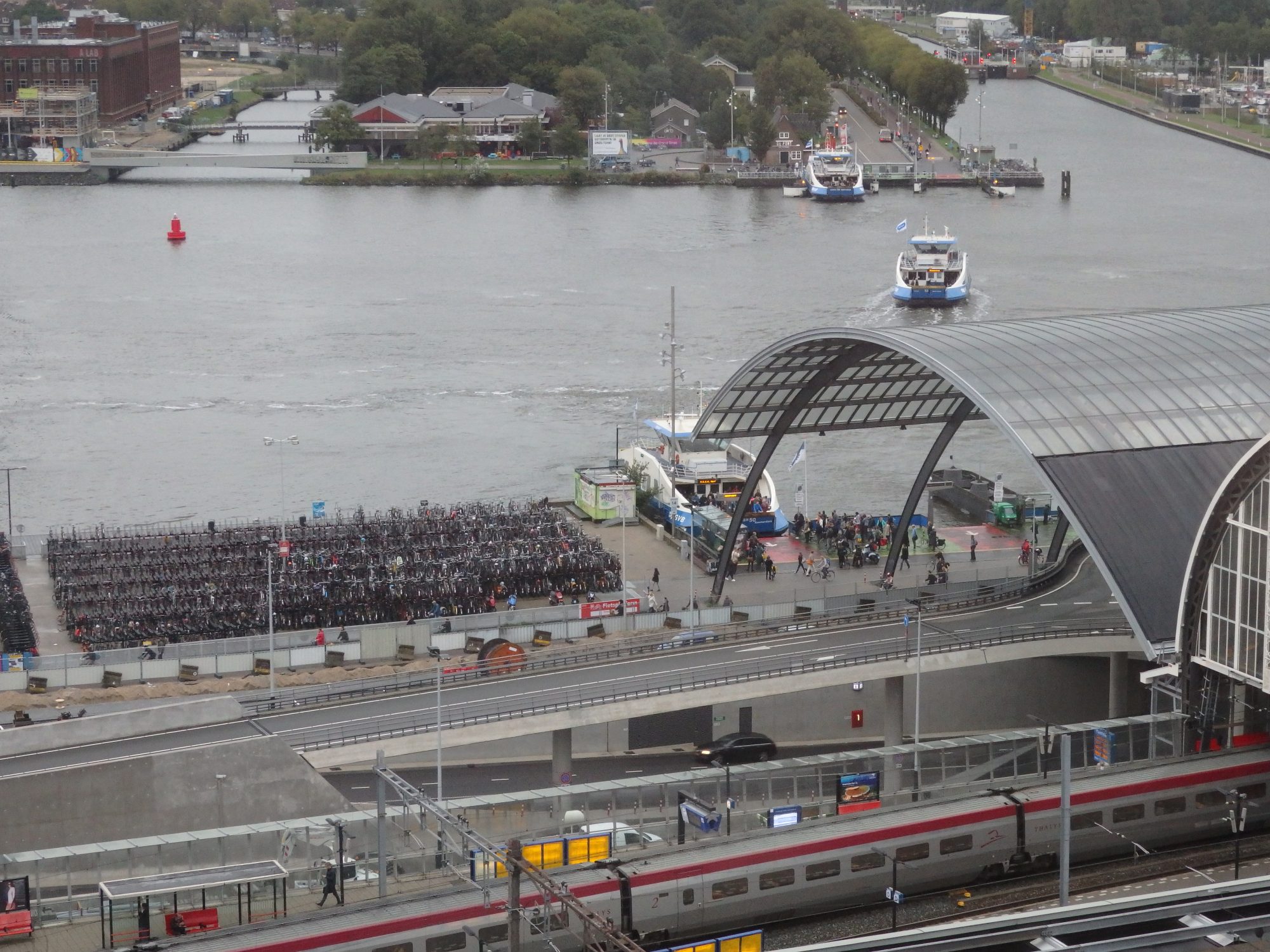 This view shows, on the right, a bit of the glass roof of the new addition to the back of Amsterdam Central Station. You can see the ferry landing and one ferry crossing the IJ river. Notice the bike parking next to the ferry landing!