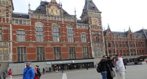 A view of the front of Amsterdam Central Station: red brick with ornate decorations along its top and a tower with a gold clock.