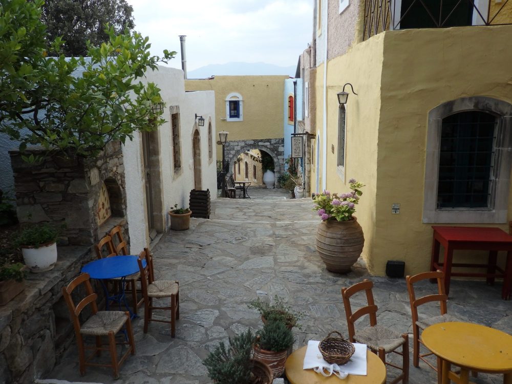 A "street" in Arolithos Traditional Cretan Village. The street is stone paved, with tables and chairs in the foreground. It slants downwards straight ahead, with some stairs down. On either side are low buildings painted white or light yellow.