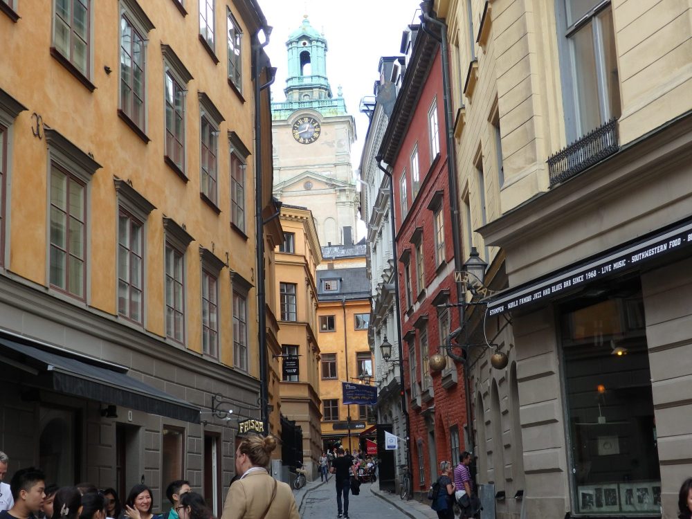 a narrow street in the oldest part of Stockholm, Gamla Stan. Older apartment buildings on both sides, and a clock tower of a church visible down the street. 