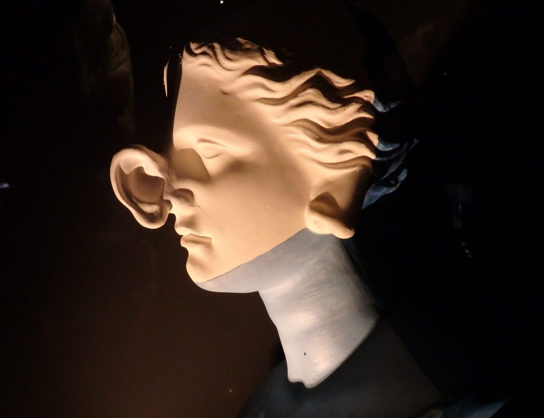 Side view of a whitish sculpture on a black background. It is a woman's head, seen from the side, and where the nose should be, an ear sticks out from her face. Where the ear should be, a nose sticks out.