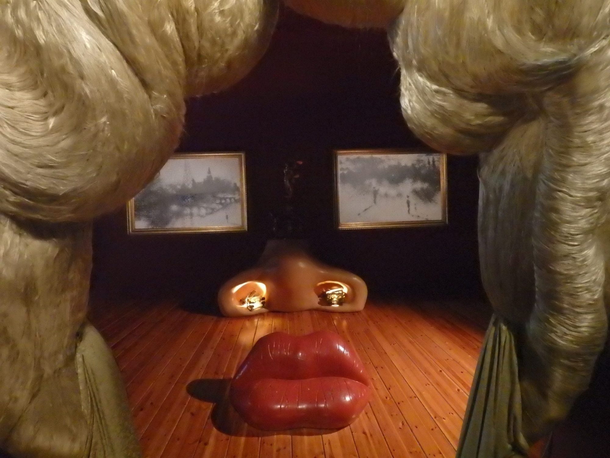 The "hair" fills the left and right sides of the photo. Ahead, on the floor is the lip-shaped sofa. Behind that, the shape of a nose, lit up from inside the nostrils. On the wall behind that: two horizontal paintings that are indistinct, but from this vantage point look like eyes.