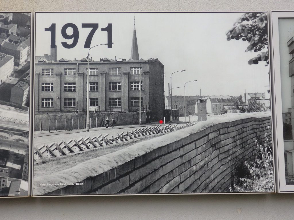 The photo has 1971 in the corner and shows the wall in the foreground, a row of large barriers to vehicles behind and parallel to the wall, and a building beyond that.