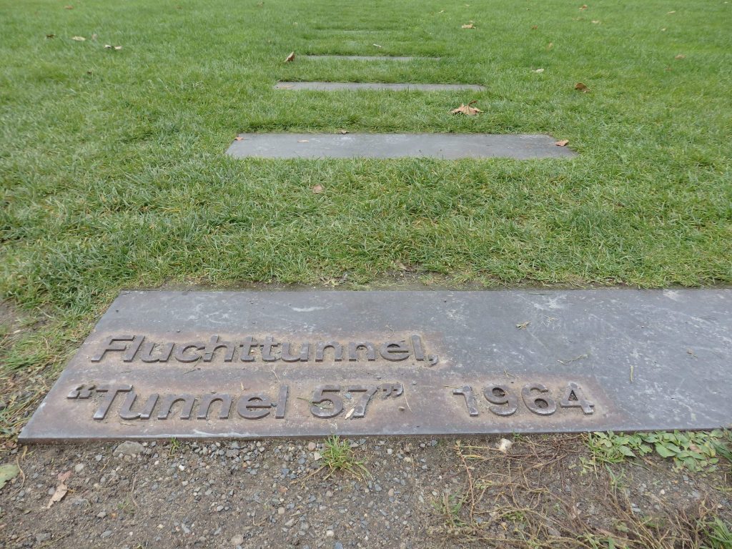 Flat metal rectangles set flat in the grass, in a row.The nearest one reads "Fluchttunnel, 'Tunnel 57" 1964.'