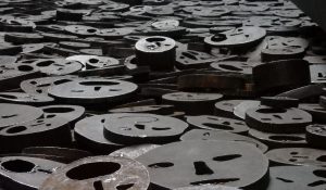 Flat metal disks, piled haphazardly, covering the floor. Each has a simple face, most of them with a round O for a mouth as if screaming.