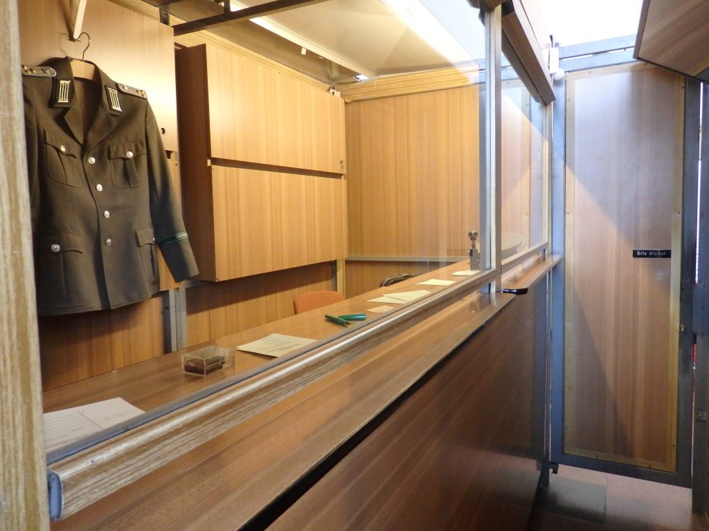 A narrow hallway with, on the left, a high counter with glass above it. A border guard uniform hangs at the back of the booth.