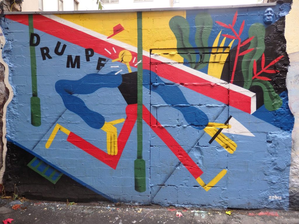 Almost a stick figure but in bright primary colors, running down a sidewalk and running straight into a poll. A word behind him reads "Drumpf."