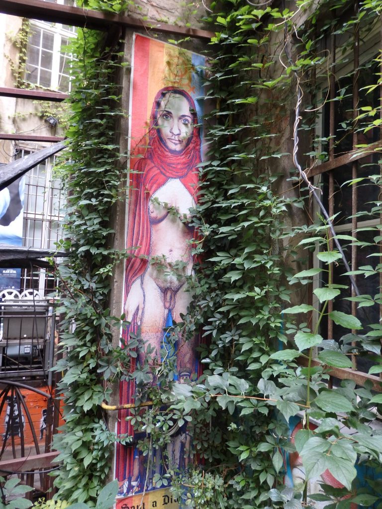 A vertical painting depicts a person with female breasts and male genitals.