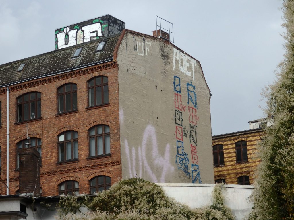 An apartment building of maybe 4 stories, with a tag on a small structure on the roof, a vertical strip of graffiti down the side wall, and a blurry smear on the same side wall.