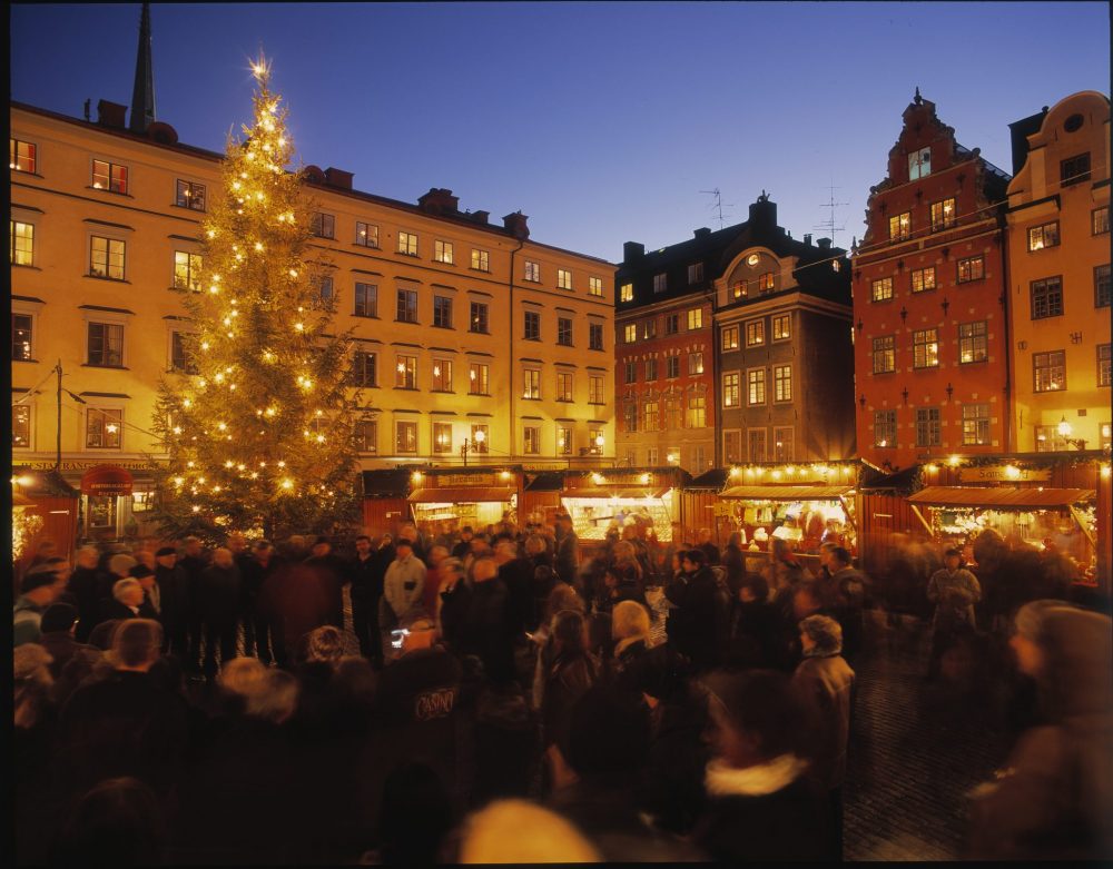 Two sides of the square are visible: old buildings, their windows lit up and the night sky behind. Below is a row of brightly-lit stalls and a crowd of people as wel as a tall Xmas tree lit with white lights. photo by Jeppe Wikström/ mediabank.visitstockholm.com 