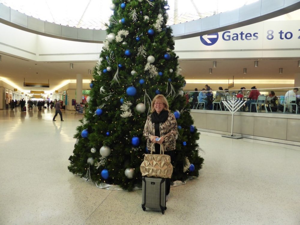 Irene poses in front of a tall Xmas tree with blue and white balls, in the middle of an open space inside an airport. She wears a warm jacket and has a grey wheeled bag and a large handbag. Behind her a sign is visible pointing to gates. Christmas in the airport. Photo courtesy of Irene S. Levine