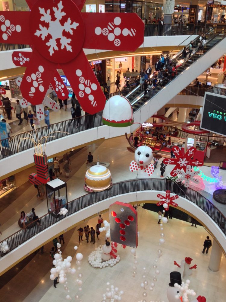 a shopping center in Chiang Mai, Thailand, looking from one of the floors into the open atrium in the middle of the mall. Oversized Xmas decorations hang in the atrium. Below, on the bottlm level, round white balls surround a snowman. Photo courtesy of Jane Dempster-Smith