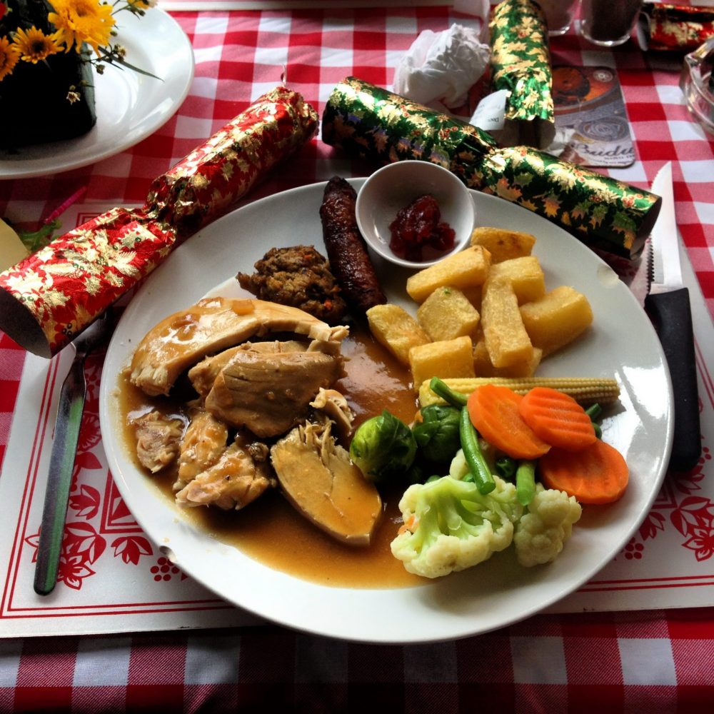 A plate of food that includes some sort of meat with brown gravy, some fries, mixed vegetables, and a few other small items I can't identify. Next to the plate are two Xmas crackers. Photo courtesy of Jane Dempster-Smith