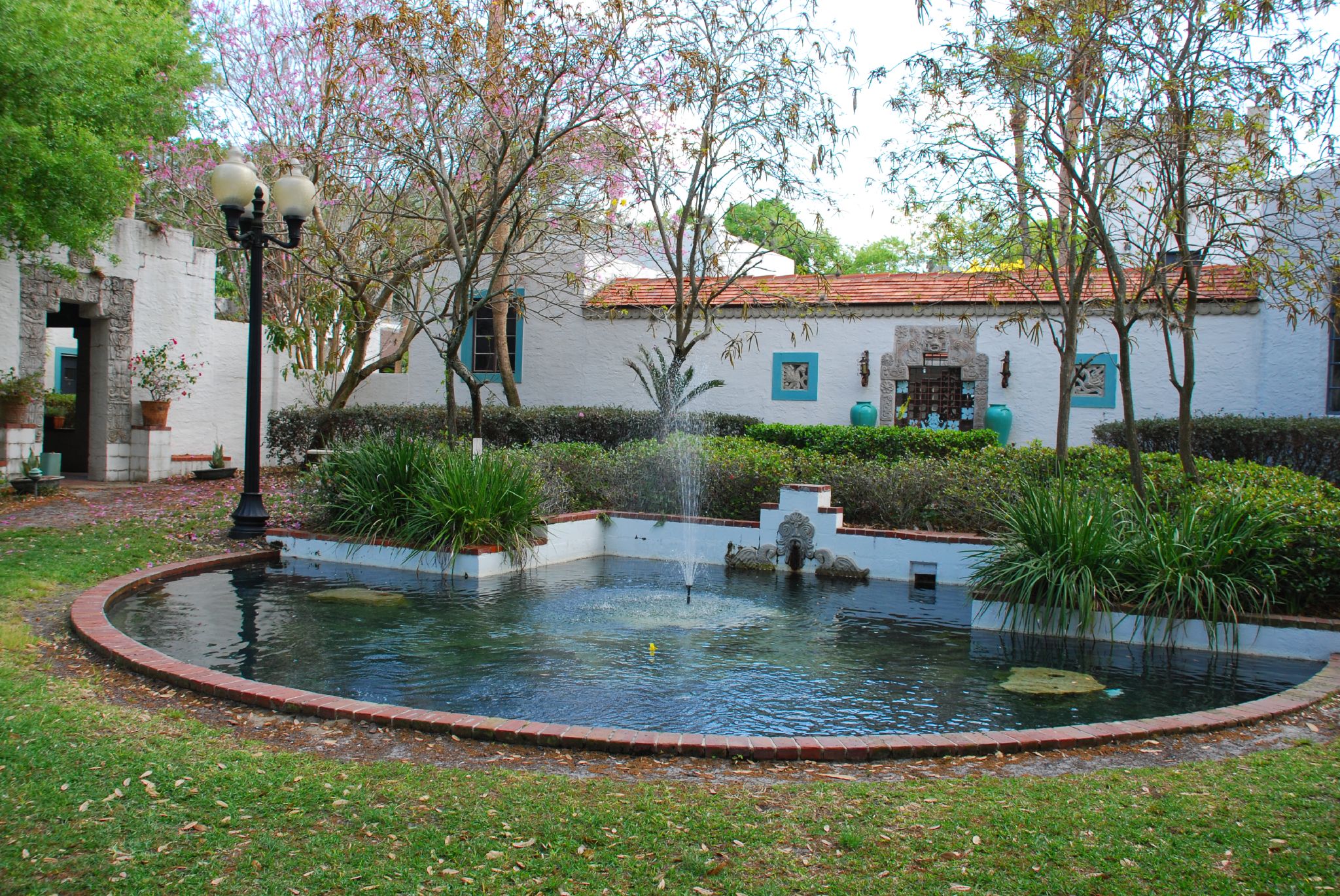A semi-circular fountain in the foreground, with water spraying straight up. Behind, the adobe museum building: single-story, whitewashed, with a red tile roof.