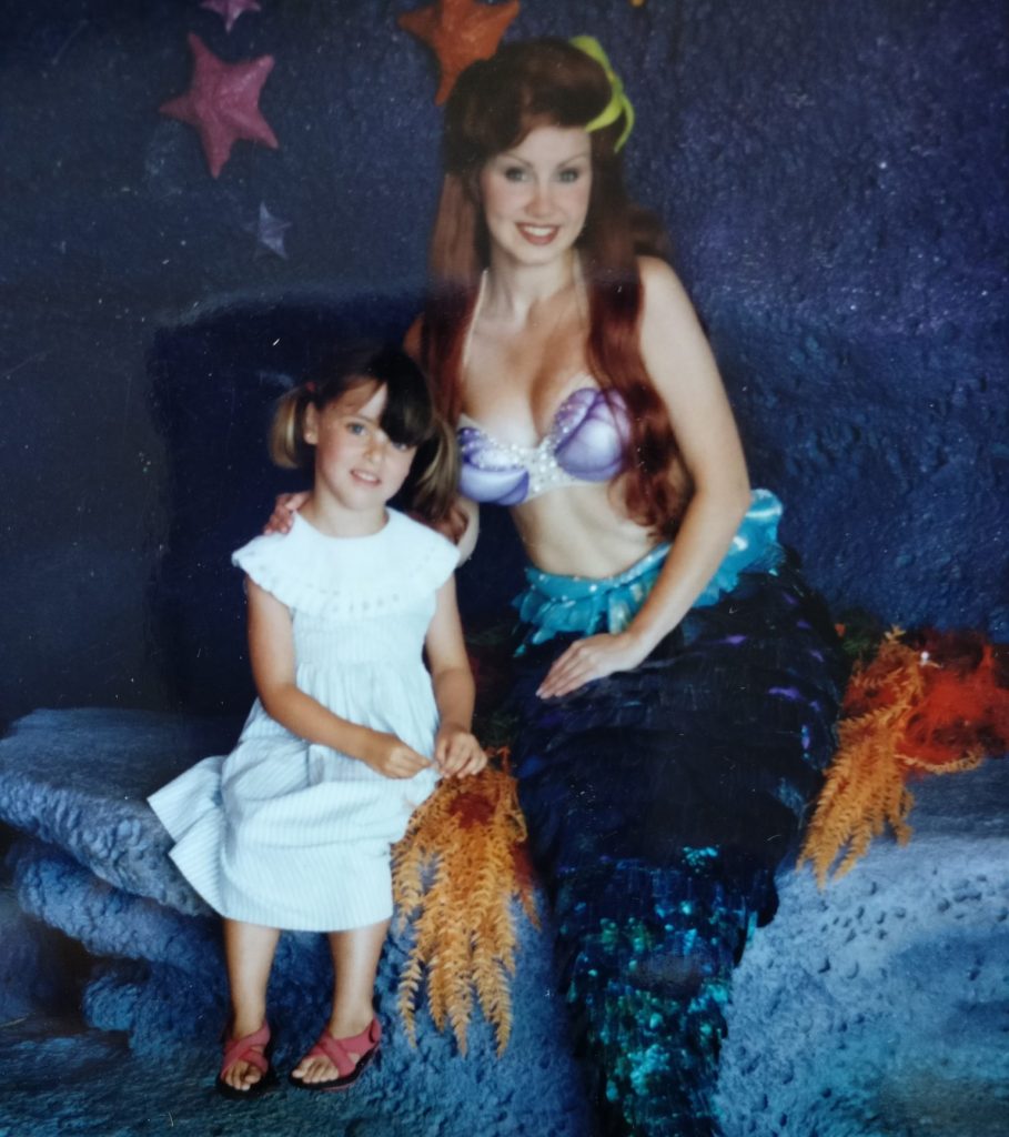 Woman dressed as Ariel: red long hair, bikini top, mermaid's tail, sitting on a stone-look bench. Next to her, a little girl wearing a light blue dress, hair in pigtails, red sandals. 