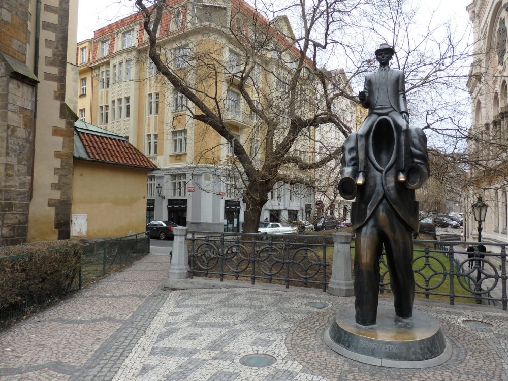 In a small square, with apartment buildings surrounding it, next to a tree, the statue depicts a man, his feet submerged in water? mud?, carrying a smaller man on his shoulders. The man sitting on his shoulders looks like a normal man, in a jacket, wearing a hat. But the man doing the carrying has no head. Instead, the collar of his jacket gapes and no chest or head are visible: just a hole. 