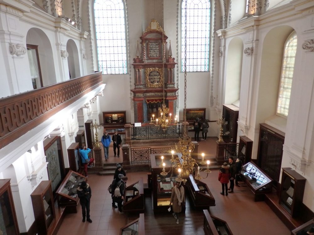 The interior of the Klausen Synagogue, one of the Synagogues in Prague, seen from upstairs in the "women's gallery." A high, baroque structure frames what would have been the torah ark in the opposite wall. On either side of it is a large, arched window, letting in a lot of light. Below, on the ground floor, various display cases line the walls and down the middle. People are standing and looking at them here and there. 