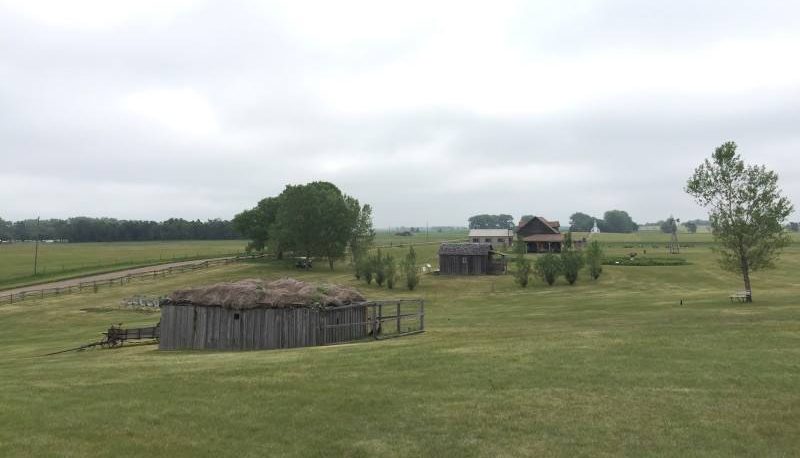 In the foreground, what looks like it might be a barn of some sort: grey wooden sides and a sod roof. Beyond it, across a flat grassy field: 3 more houses. One looks like a one-room wooden house. The other two look more substantial: one painted white, single story; one brown one with two stories and a veranda.