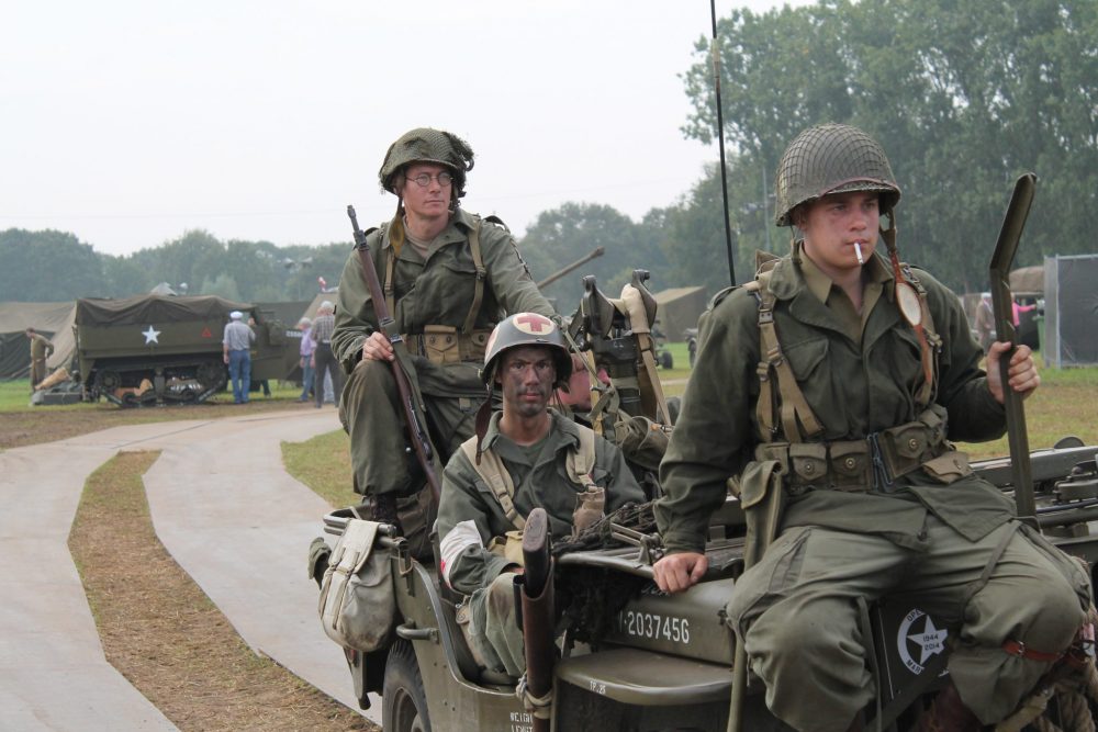 Several men in khaki camoflage uniforms, wearing helmets and holding rifles, sit on a jeep. 