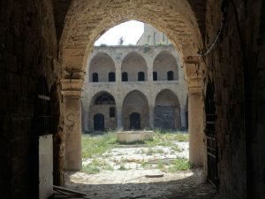 Seen through its entry arch, the khan is abandoned and its courtyard is weedy. On the opposite side, archways on the ground floor, more archways on the upper floor shading a balcony, doors visible behind that.