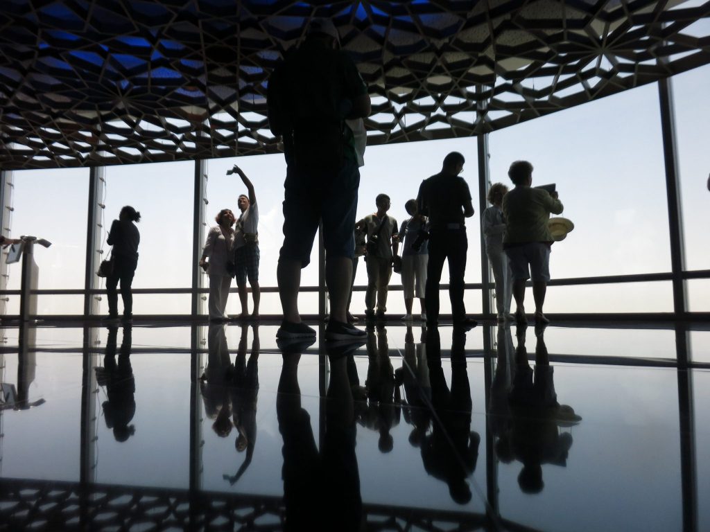 Tourists in silhouette stand on a shiny black floor and look out floor-to-ceiling windows or stand with their backs to the windows taking selfies.